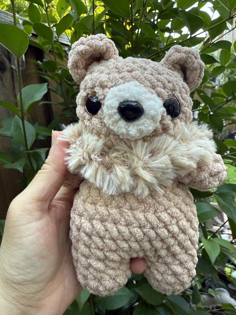 Peut inclure: Un ours en crochet marron clair avec un museau blanc duveteux et un nez noir. L'ours a de grands yeux noirs et de petites oreilles arrondies. L'ours est tenu dans la main d'une personne sur un fond de feuilles vertes.
