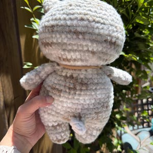 Pu&ograve; includere: Una bambola a forma di gatto all'uncinetto fatta a mano nei toni del grigio e del marrone. Il peluche ha una testa rotonda con piccole orecchie, un corpo cilindrico e braccia e gambe minuscole. Una fascia marrone chiaro &egrave; intorno al collo. La coda del gatto &egrave; visibile.