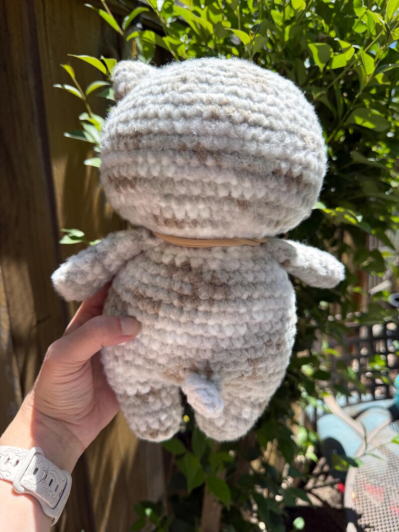 Pu&ograve; includere: Una bambola a forma di gatto all'uncinetto fatta a mano nei toni del grigio e del marrone. Il peluche ha una testa rotonda con piccole orecchie, un corpo cilindrico e braccia e gambe minuscole. Una fascia marrone chiaro &egrave; intorno al collo. La coda del gatto &egrave; visibile.