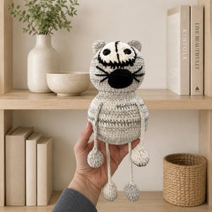May include: A handmade crochet cat toy featuring a round, mottled grey and white yarn body with a large, oval-shaped black nose, embroidered black whiskers, and two round eyes with black pupils. The cat has long, thin dangling legs and arms.