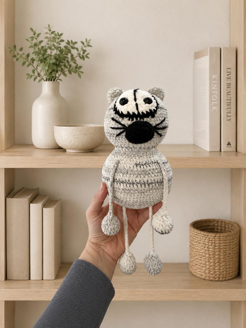 May include: A handmade crochet cat toy featuring a round, mottled grey and white yarn body with a large, oval-shaped black nose, embroidered black whiskers, and two round eyes with black pupils. The cat has long, thin dangling legs and arms.