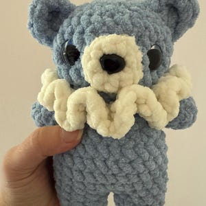 May include: A crocheted blue teddy bear with a white crocheted collar. The bear has large black eyes and a black nose.