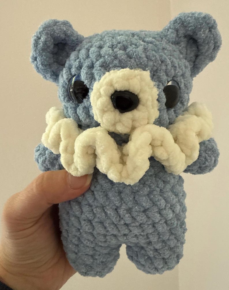 May include: A crocheted blue teddy bear with a white crocheted collar. The bear has large black eyes and a black nose.