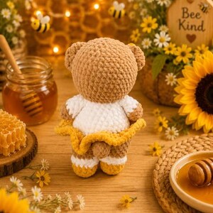 K&ouml;nnte beinhalten: Ein geh&auml;kelter Teddyb&auml;r aus hellbraunem Garn mit einem wei&szlig;en Oberteil und einem gelben Rock. Der B&auml;r hat abgerundete Ohren und einen kleinen Schwanz, der auf der R&uuml;ckseite sichtbar ist. Die Garnstruktur ist weich und flauschig verarbeitet.
