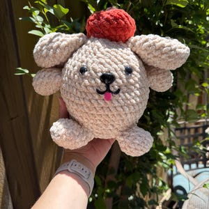以下が含まれることがあります： 赤いポンポン帽子をかぶった、手作りのベージュのかぎ針編みの犬。犬は黒い目、黒い鼻、ピンクの舌をしています。ぬいぐるみは丸く、垂れ耳と小さな手足が付いています。