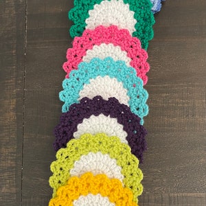 Peut inclure: Une pile de huit dessous-de-verre en crochet de diff&eacute;rentes couleurs, notamment bleu, vert, rose, turquoise, violet, jaune et blanc. Les dessous-de-verre sont dispos&eacute;s en arc-en-ciel, le bleu en haut et le jaune en bas.
