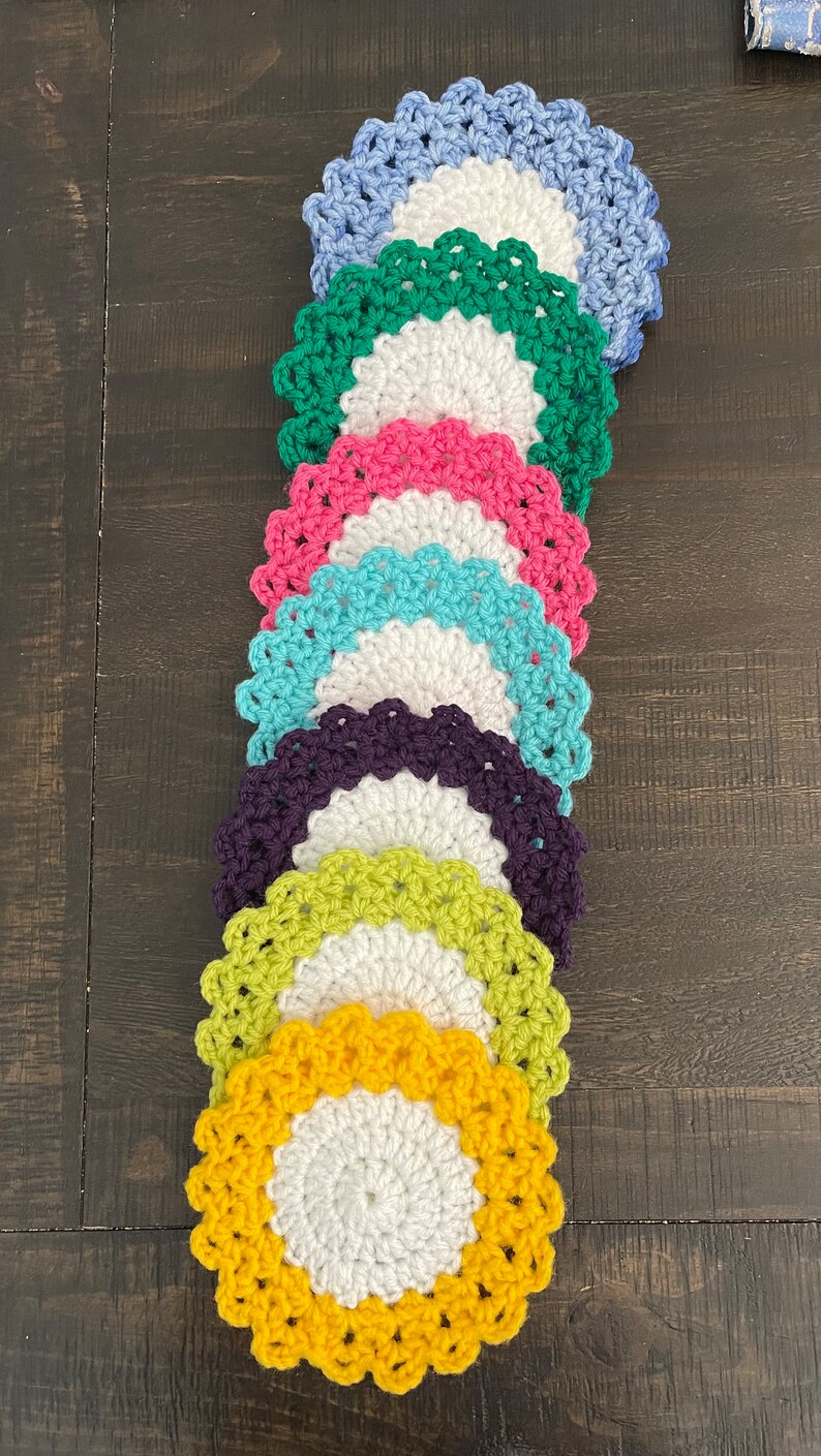 Peut inclure: Une pile de huit dessous-de-verre en crochet de diff&eacute;rentes couleurs, notamment bleu, vert, rose, turquoise, violet, jaune et blanc. Les dessous-de-verre sont dispos&eacute;s en arc-en-ciel, le bleu en haut et le jaune en bas.