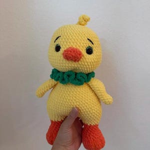 Op de afbeelding: Een handgemaakt gehaakt geel kuiken met oranje voeten en snavel, en een groene kraag. Het kuiken heeft zwarte ogen en een kleine pluk geel garen op zijn hoofd. Een schattig, zacht en knuffelbaar speelgoed.