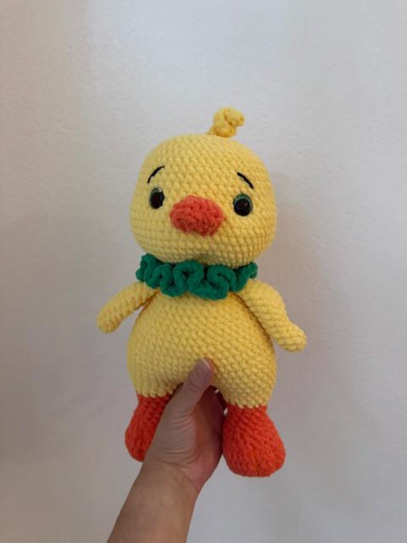 Op de afbeelding: Een handgemaakt gehaakt geel kuiken met oranje voeten en snavel, en een groene kraag. Het kuiken heeft zwarte ogen en een kleine pluk geel garen op zijn hoofd. Een schattig, zacht en knuffelbaar speelgoed.