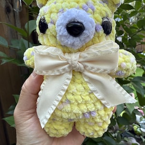 May include: A handmade yellow crochet bear with purple speckles, black eyes, and a cream-colored bow. The bear is soft and plush, perfect for a child's toy or nursery decor.