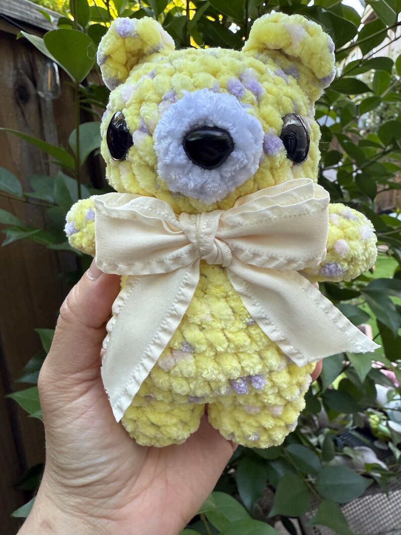 May include: A handmade yellow crochet bear with purple speckles, black eyes, and a cream-colored bow. The bear is soft and plush, perfect for a child's toy or nursery decor.