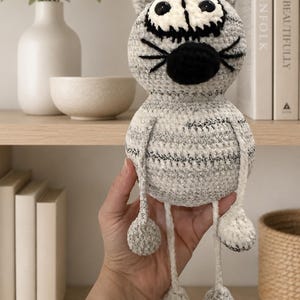 May include: A crocheted cat plush toy with a round head and body made of variegated gray and white yarn. It features large black plastic eyes, a black stitched nose, and black whiskers. The toy has long, thin dangling arms and legs with round feet.