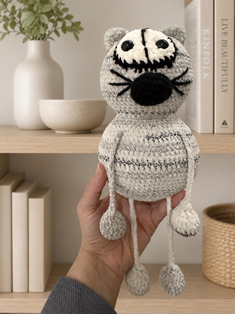 May include: A crocheted cat plush toy with a round head and body made of variegated gray and white yarn. It features large black plastic eyes, a black stitched nose, and black whiskers. The toy has long, thin dangling arms and legs with round feet.