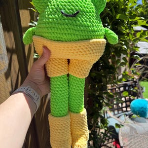 Op de afbeelding: Een handgemaakt, gehaakt kikkerknuffel in heldergroen, geel en zwart. De kikker heeft grote, ronde ogen, een lachende mond en draagt gele shorts en laarzen. Het speelgoed is gemaakt van zacht, getextureerd garen.