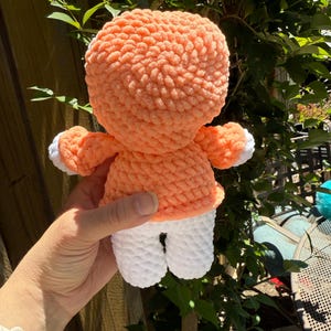 May include: A handmade crochet doll with a peach-coloured head and torso, white legs, and small arms. The doll has a textured, plush appearance. The doll is held in front of a green leafy background.
