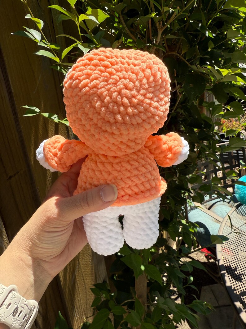 May include: A handmade crochet doll with a peach-coloured head and torso, white legs, and small arms. The doll has a textured, plush appearance. The doll is held in front of a green leafy background.