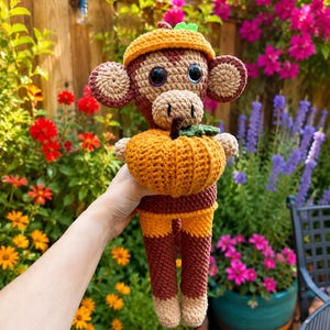 May include: A crocheted monkey toy featuring dark brown and tan yarn, wearing a small orange hat and holding a textured orange pumpkin with a green stem. The monkey has large black plastic eyes, round ears, and a long tail, crafted with detailed stitching.