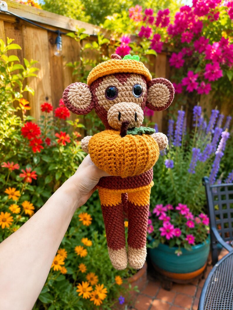 May include: A crocheted monkey toy featuring dark brown and tan yarn, wearing a small orange hat and holding a textured orange pumpkin with a green stem. The monkey has large black plastic eyes, round ears, and a long tail, crafted with detailed stitching.