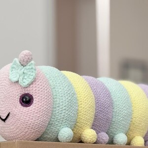以下が含まれることがあります： パステルカラーの手編みのかぎ針編みのイモムシ。頭はピンク色で、黒い目、笑顔の口、ミントグリーンのリボンが付いています。体はミントグリーン、イエロー、ラベンダーのセグメントが交互に並んでいます。各セグメントの基部には小さなポンポンが付いています。