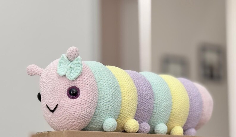 以下が含まれることがあります： パステルカラーの手編みのかぎ針編みのイモムシ。頭はピンク色で、黒い目、笑顔の口、ミントグリーンのリボンが付いています。体はミントグリーン、イエロー、ラベンダーのセグメントが交互に並んでいます。各セグメントの基部には小さなポンポンが付いています。