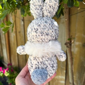 Peut inclure: Un lapin en peluche fait &agrave; la main, avec un corps duveteux mouchet&eacute; de blanc et de gris, de longues oreilles et une petite queue en pompon bleu. Le lapin a un col en fourrure blanche. Le jouet est tenu dans une main.
