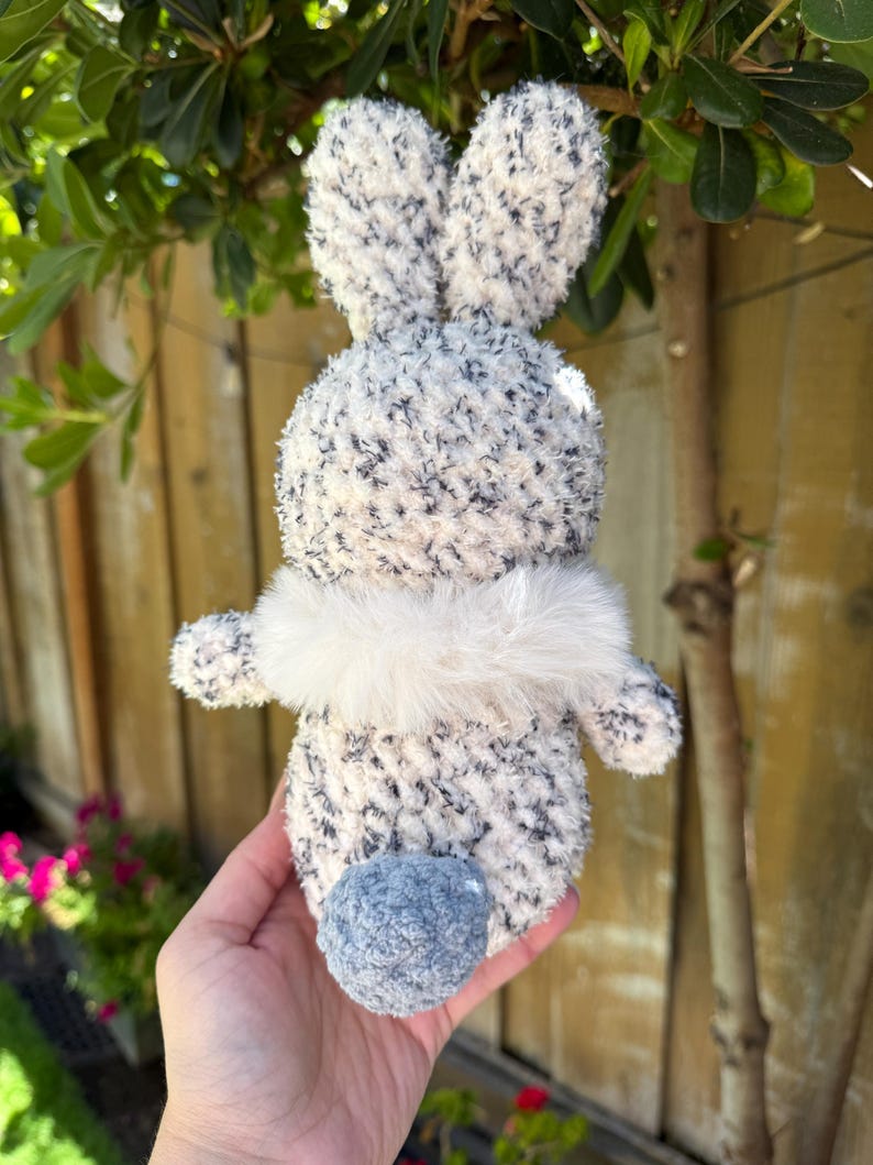 Peut inclure: Un lapin en peluche fait &agrave; la main, avec un corps duveteux mouchet&eacute; de blanc et de gris, de longues oreilles et une petite queue en pompon bleu. Le lapin a un col en fourrure blanche. Le jouet est tenu dans une main.