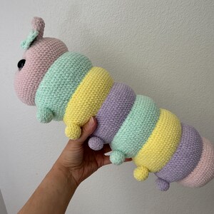 以下が含まれることがあります： パステルカラーの毛糸で作られた、手編みのカラフルなイモムシのぬいぐるみ。ピンク、ミントグリーン、イエロー、パープルを使用。黒い目と小さなポンポンが付いています。手作りの柔らかいおもちゃです。