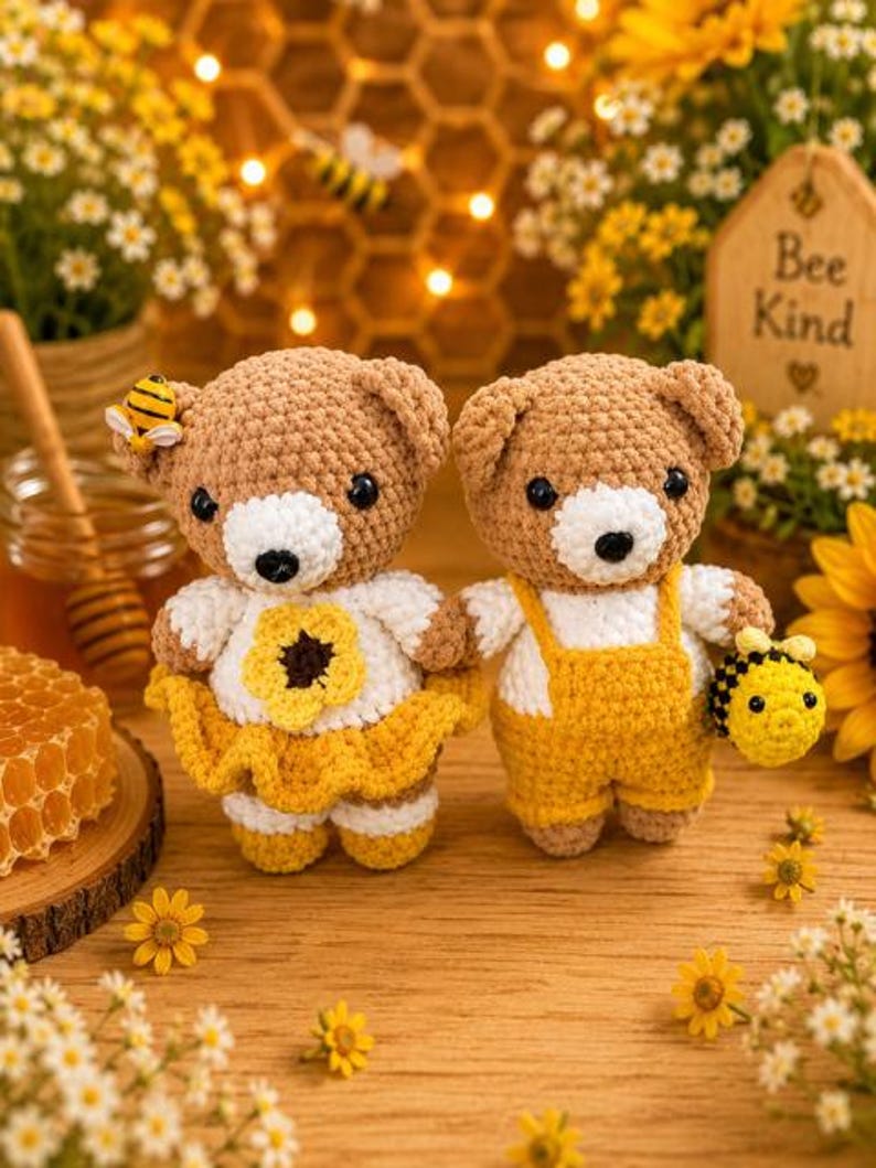 K&ouml;nnte beinhalten: Zwei geh&auml;kelte Teddyb&auml;ren mit hellbraunen Garnk&ouml;rpern und wei&szlig;en Schnauzen. Ein B&auml;r tr&auml;gt ein wei&szlig;es Oberteil mit einer gelben Blume und einem gelben Rock sowie einer kleinen Biene am Ohr. Der andere B&auml;r tr&auml;gt ein wei&szlig;es Oberteil mit gelber Latzhose und h&auml;lt eine kleine geh&auml;kelte Biene.