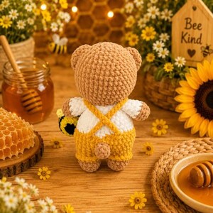 K&ouml;nnte beinhalten: Ein geh&auml;kelter Teddyb&auml;r aus hellbraunem Garn, der ein wei&szlig;es Hemd und eine gelbe Latzhose mit gekreuzten Tr&auml;gern tr&auml;gt. Der B&auml;r hat einen kleinen runden Schwanz und h&auml;lt ein kleines geh&auml;keltes Bienenspielzeug. Der B&auml;r ist von hinten zu sehen.