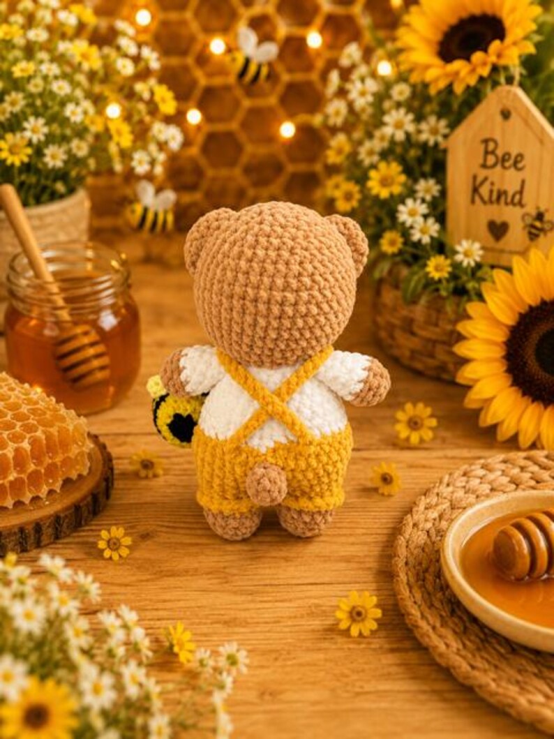 K&ouml;nnte beinhalten: Ein geh&auml;kelter Teddyb&auml;r aus hellbraunem Garn, der ein wei&szlig;es Hemd und eine gelbe Latzhose mit gekreuzten Tr&auml;gern tr&auml;gt. Der B&auml;r hat einen kleinen runden Schwanz und h&auml;lt ein kleines geh&auml;keltes Bienenspielzeug. Der B&auml;r ist von hinten zu sehen.
