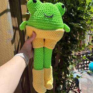 Op de afbeelding: Een handgemaakte gehaakte kikkerpop met een felgroene kop en poten, gele shorts en laarzen. De kikker heeft zwarte ogen en een lachende mond. De pop wordt vastgehouden tegen een houten hek en groen gebladerte.