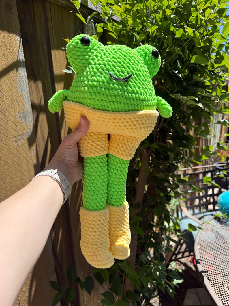 Op de afbeelding: Een handgemaakte gehaakte kikkerpop met een felgroene kop en poten, gele shorts en laarzen. De kikker heeft zwarte ogen en een lachende mond. De pop wordt vastgehouden tegen een houten hek en groen gebladerte.
