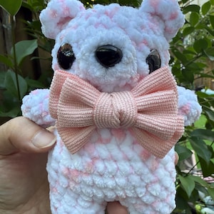 May include: A handmade, plush teddy bear with a white and pink speckled pattern. The bear has large black eyes and a light pink bow tie. The bear is being held in a person's hand, with green foliage in the background.