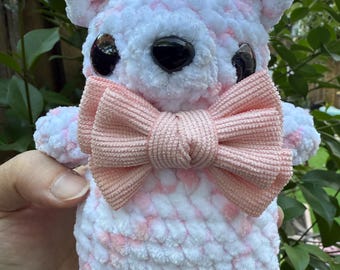 Peut inclure: Un ours en peluche fait à la main avec un motif moucheté blanc et rose. L'ours a de grands yeux noirs et un nœud papillon rose clair. L'ours est tenu dans la main d'une personne, avec un feuillage vert en arrière-plan.