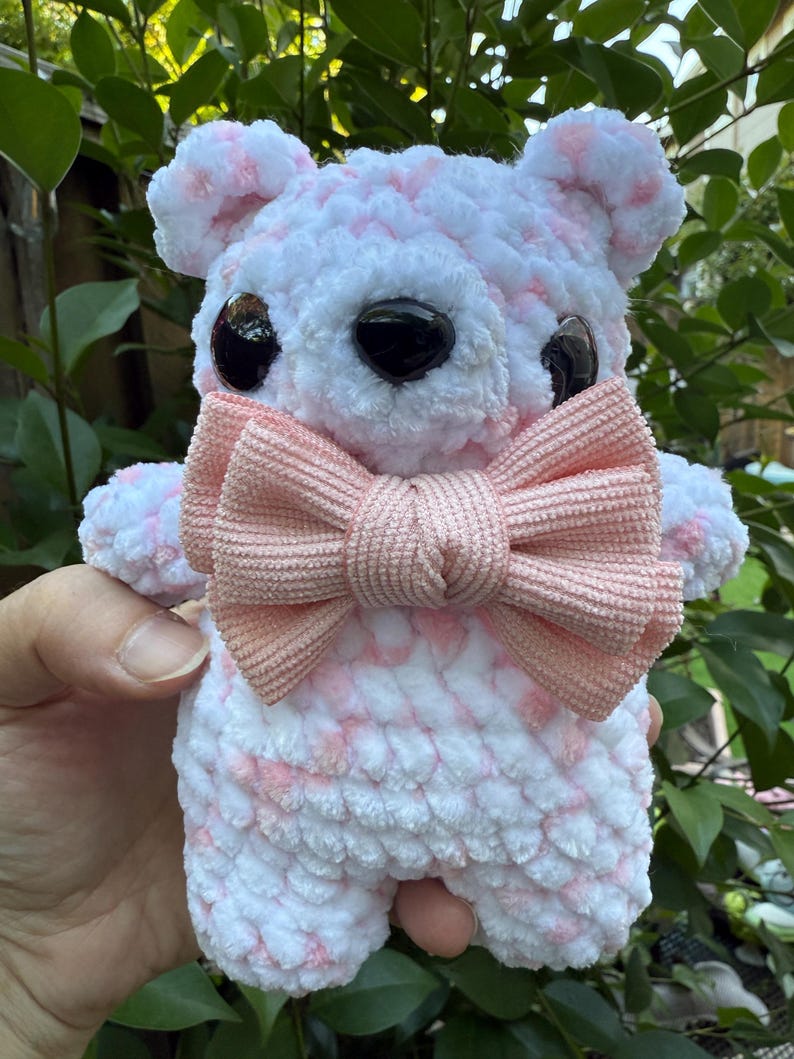 May include: A handmade, plush teddy bear with a white and pink speckled pattern. The bear has large black eyes and a light pink bow tie. The bear is being held in a person's hand, with green foliage in the background.