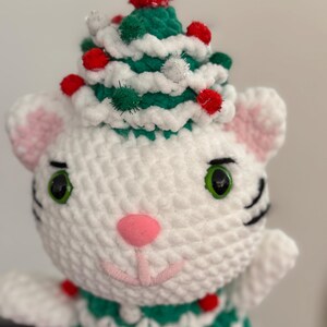 Pode incluir: Boneca de gato de croch&ecirc; branco feita &agrave; m&atilde;o usando um chap&eacute;u de &aacute;rvore de Natal verde e branco adornado com pompons vermelhos. O gato tem olhos verdes, nariz rosa e orelhas internas rosas. O corpo do gato tamb&eacute;m &eacute; decorado com pompons vermelhos.