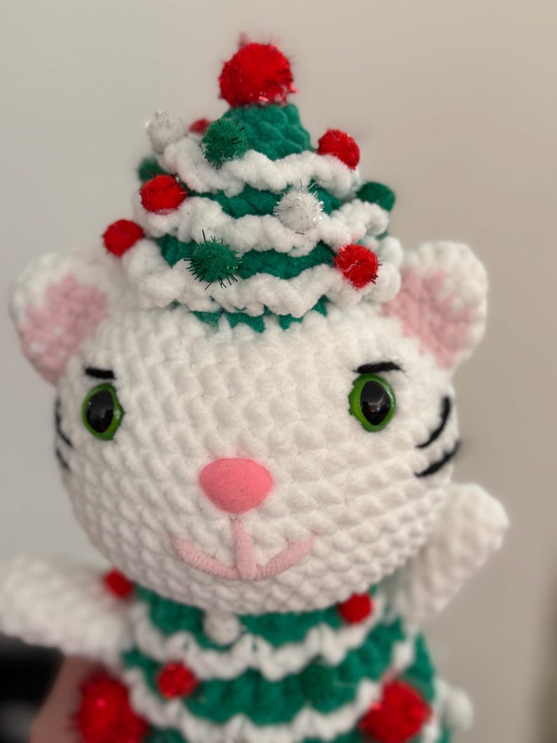 Pode incluir: Boneca de gato de croch&ecirc; branco feita &agrave; m&atilde;o usando um chap&eacute;u de &aacute;rvore de Natal verde e branco adornado com pompons vermelhos. O gato tem olhos verdes, nariz rosa e orelhas internas rosas. O corpo do gato tamb&eacute;m &eacute; decorado com pompons vermelhos.