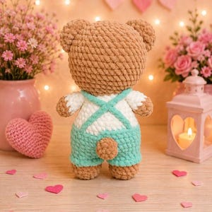 K&ouml;nnte beinhalten: Ein geh&auml;kelter Pl&uuml;sch-Teddyb&auml;r aus hellbraunem Garn, der eine wei&szlig;e und mintgr&uuml;ne Latzhose mit &uuml;berkreuzten Tr&auml;gern auf dem R&uuml;cken tr&auml;gt. Der B&auml;r hat einen kleinen runden Schwanz und eine weiche, strukturierte Oberfl&auml;che und steht mit dem R&uuml;cken zum Betrachter.