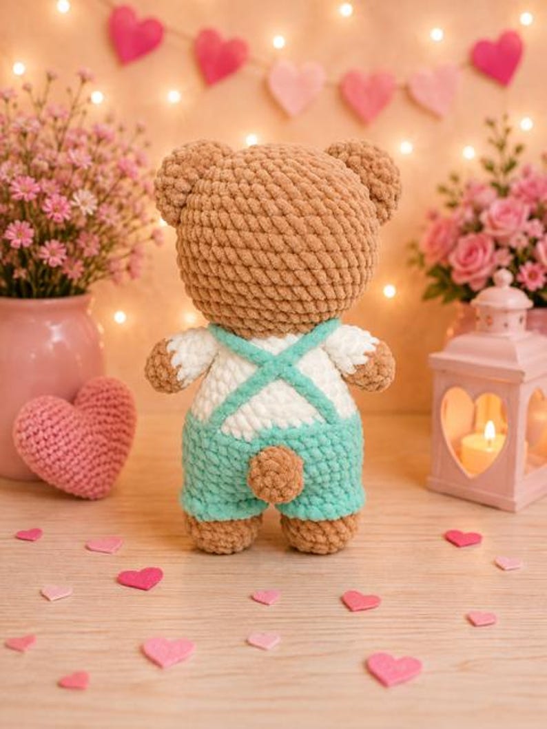 K&ouml;nnte beinhalten: Ein geh&auml;kelter Pl&uuml;sch-Teddyb&auml;r aus hellbraunem Garn, der eine wei&szlig;e und mintgr&uuml;ne Latzhose mit &uuml;berkreuzten Tr&auml;gern auf dem R&uuml;cken tr&auml;gt. Der B&auml;r hat einen kleinen runden Schwanz und eine weiche, strukturierte Oberfl&auml;che und steht mit dem R&uuml;cken zum Betrachter.