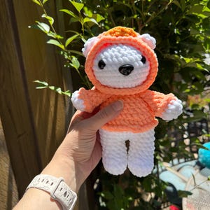 May include: A handmade crochet bear doll wearing an orange hoodie. The bear is white with a black nose and eyes. The hoodie has a matching orange hood and sleeves. The doll is held in front of a green leafy background.