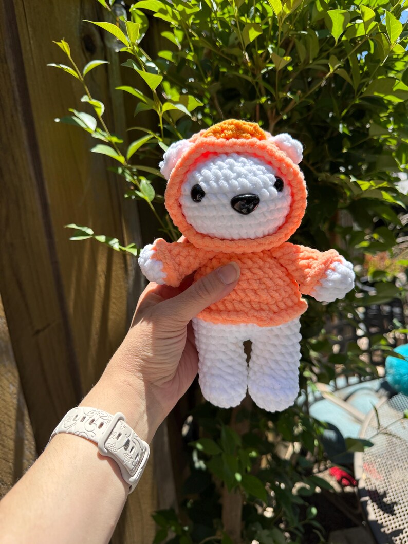 May include: A handmade crochet bear doll wearing an orange hoodie. The bear is white with a black nose and eyes. The hoodie has a matching orange hood and sleeves. The doll is held in front of a green leafy background.