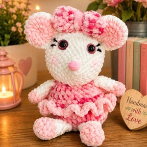 May include: A plush crochet mouse toy featuring white and pink yarn, large round ears, a pink nose, and a matching pink bow on its head. The mouse wears a ruffled pink and white dress and has black plastic eyes with stitched eyelashes.