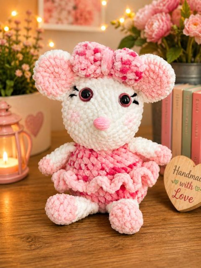 May include: A plush crochet mouse toy featuring white and pink yarn, large round ears, a pink nose, and a matching pink bow on its head. The mouse wears a ruffled pink and white dress and has black plastic eyes with stitched eyelashes.