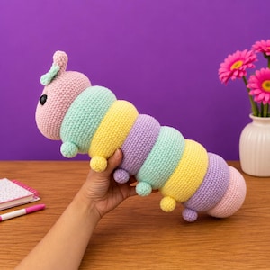 以下が含まれることがあります： ピンク、ミントグリーン、イエロー、ラベンダーのパステルカラーの手作りの編み物で作られた毛虫のおもちゃ。 ぬいぐるみのような毛虫は、黒い目と小さな触角を持つ丸い頭をしています。 おもちゃは紫色の背景に対して保持されています。