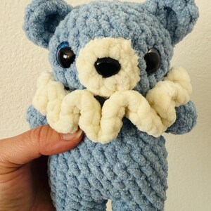 May include: A blue crocheted teddy bear with white crocheted details around the neck and large black eyes.