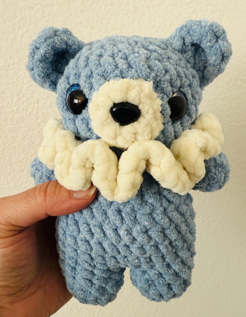 May include: A blue crocheted teddy bear with white crocheted details around the neck and large black eyes.
