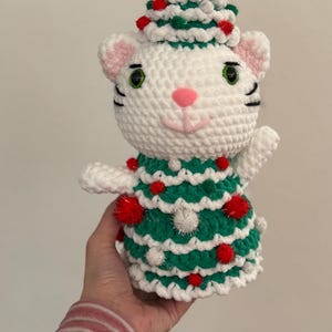 Pode incluir: Figura de gato de croch&ecirc; branco feita &agrave; m&atilde;o, vestindo um vestido e chap&eacute;u de &aacute;rvore de Natal verde e branco. O gato tem listras pretas, olhos verdes e nariz rosa. Pompons vermelhos e brancos decoram a &aacute;rvore.