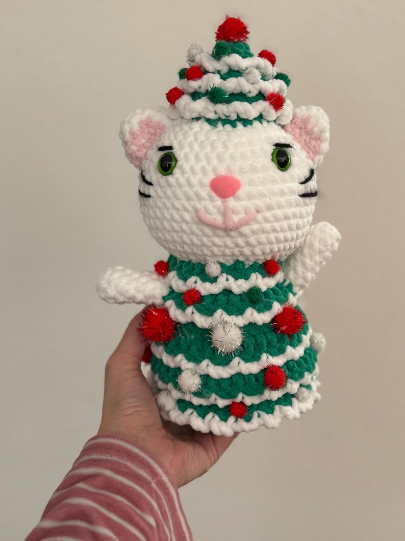 Pode incluir: Figura de gato de croch&ecirc; branco feita &agrave; m&atilde;o, vestindo um vestido e chap&eacute;u de &aacute;rvore de Natal verde e branco. O gato tem listras pretas, olhos verdes e nariz rosa. Pompons vermelhos e brancos decoram a &aacute;rvore.