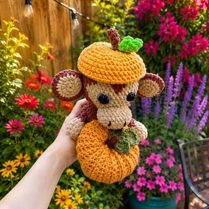 May include: A crocheted monkey plush toy wearing an orange pumpkin-shaped hat and holding a small orange pumpkin with a green leaf. The monkey has brown and beige yarn, black plastic eyes, and round ears, crafted with a textured stitch pattern.