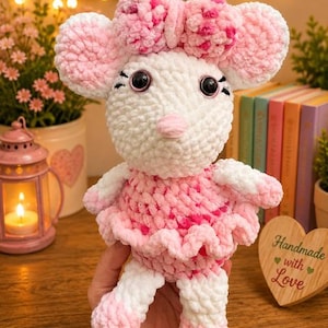 May include: A plush crochet mouse toy made of soft white and pink yarn. The mouse features large round ears, a pink nose, black plastic eyes, and a pink and white patterned bow on its head. It wears a ruffled pink and white skirt and has matching pink and white feet.